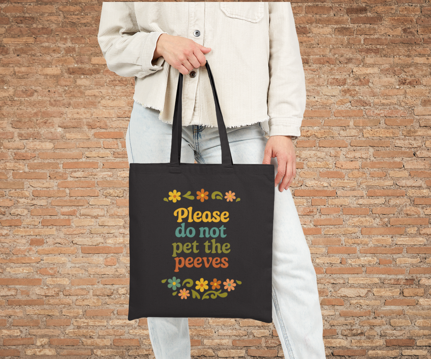 Person holding a black tote bag with colorful text please do not pet the peeves and flower graphics against a brick wall.