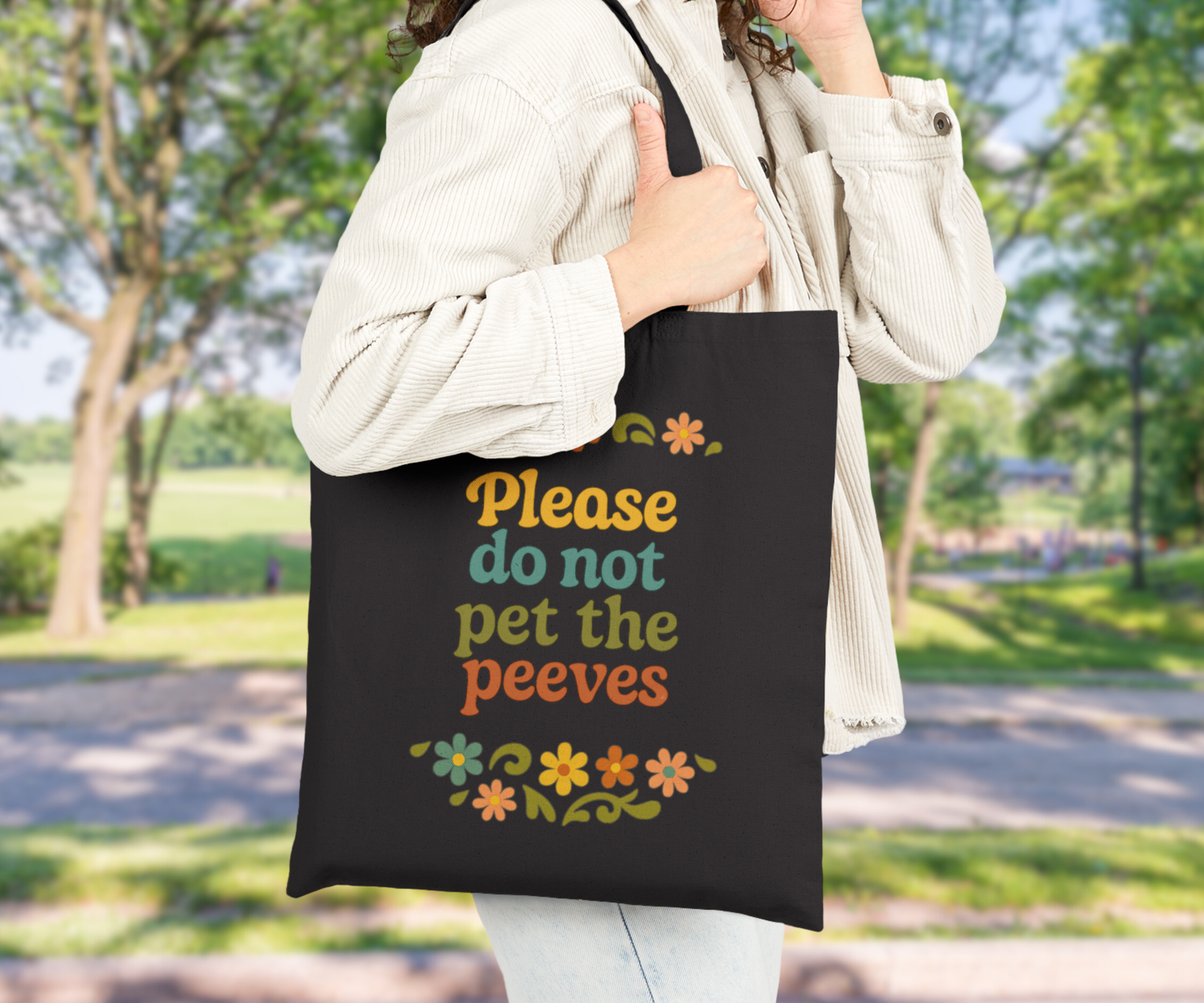 Person holding a black tote bag with colorful text please do not pet the peeves and floral designs outdoors.